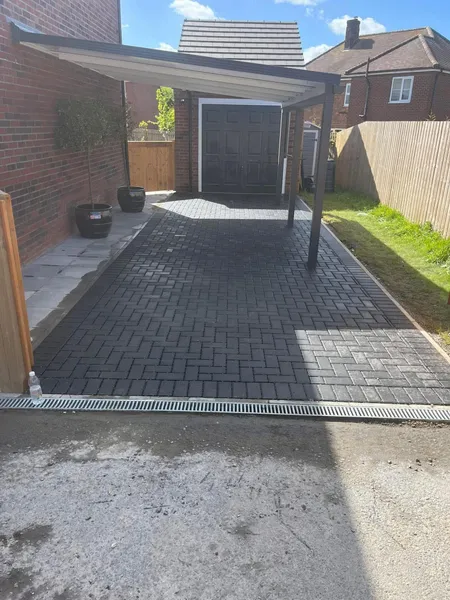 Charcoal block paving drive under a carport canopy with a channel drain across the front