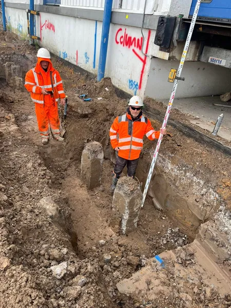 Two workers in a commercial trench with a survey staff beside a warehouse wall