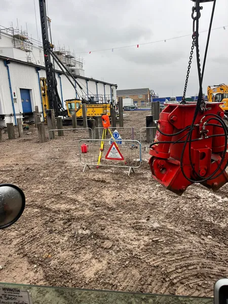 Red pile cropper hanging over a muddy site with a piling rig and barriers behind