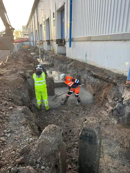 Workers levelling wet concrete in a foundation trench beside a warehouse wall